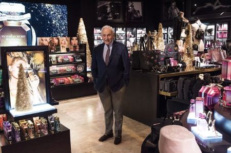 leslie wexner, the owner of Victoria Secrets stands in the middle of the VS store in a blue blazer and brown pants 